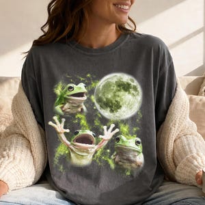 Può includere: T-shirt grigio scuro con un design stravagante di tre rane verdi e una luna verde brillante. Le rane sono raffigurate con espressioni e pose espressive, su uno sfondo di forme nebulose verdi. Una divertente maglietta grafica a tema natura.