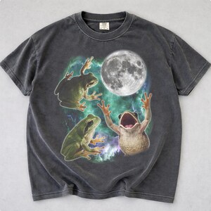 Può includere: T-shirt grigio scuro con un design stravagante di tre rane, una luna piena e uno sfondo di nebulosa. Le rane sono in varie pose, con una rana a bocca aperta e le braccia alzate. Una maglietta grafica unica.