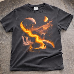 Puede incluir: Camiseta gris oscuro con una ilustración surrealista de manatíes, cascadas y una luna. El diseño incluye un resplandor naranja y amarillo ardiente que emana de los manatíes, creando un paisaje onírico. La camiseta está hecha de un material suave y cómodo.