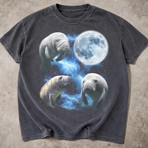 Puede incluir: Camiseta gris oscuro con un diseño de tres manatíes y una luna llena sobre un fondo de nebulosa azul y blanca. Los manatíes están representados de forma realista. La camiseta tiene cuello redondo clásico y mangas cortas.