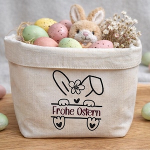 Könnte beinhalten: Ein beiger Stoff-Osterkorb mit einem Hasen-Motiv und dem Schriftzug "Frohe Ostern". Der Korb ist mit gesprenkelten Pastell-Eiern, einem Plüschhasen und Trockenblumen gefüllt. Zusätzliche Eier sind verstreut.