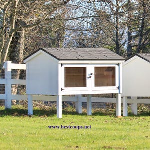 May include: Three white rabbit hutches with dark grey shingled roofs and clear windows stand on white legs in a grassy field. A white fence is in the background. The website address www.bestcoops.net is visible at the bottom.