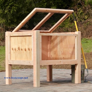 May include: A wooden raised garden bed with a hinged, screened lid. The bed has a natural wood finish and features a wire mesh panel. A yellow-handled garden tool leans against the side. The website address www.bestcoops.net is visible.