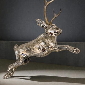 Könnte beinhalten: Eine dekorative Skulptur eines springenden Hasen, bedeckt mit Spiegelfliesen. Der Hase hat goldene Geweihe und Ohren mit einer reflektierenden Oberfläche, die das Licht einfängt. Die Skulptur steht vor einem neutralen Hintergrund und erzeugt einen auffälligen visuellen Kontrast.