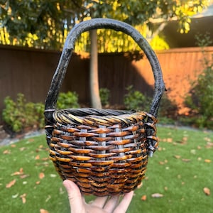 May include: A round woven basket featuring a dark brown finish and a thick, curved handle made from a natural branch. The basket body is constructed with horizontal and vertical woven strips, creating a textured, deep bowl shape with a sturdy rim.