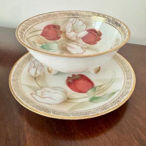 May include: A porcelain bowl and matching saucer set featuring a hand-painted tulip motif in shades of red, white, and green. Both pieces are finished with a gold-toned geometric border along the rim and the bowl rests on three small feet.