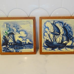 May include: Two square ceramic tiles framed in light brown wood. The left tile features a blue and white illustration of a windmill near water. The right tile displays a blue and white illustration of a sailing ship on the water with clouds.