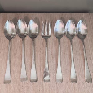 May include: A set of seven silver-toned metal utensils arranged in a row, consisting of six oval-shaped spoons and one three-pronged fork. Each piece features a smooth, polished finish with a simple, slender handle that flares slightly at the base.