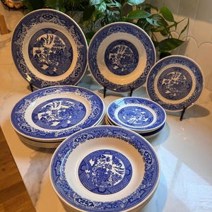 May include: A collection of white ceramic dinnerware featuring a blue willow pattern. The set includes multiple dinner plates, soup bowls, and smaller plates, all decorated with intricate blue illustrations of pagodas, trees, and birds.
