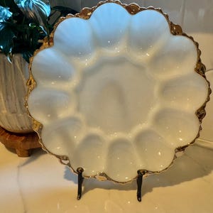May include: White, scalloped egg serving dish with a gold rim. The dish has 12 egg indentations and is supported by a black metal stand. The dish is displayed on a white surface.