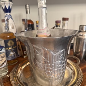 May include: A silver champagne bucket with a grape vine design, holding a bottle of sparkling wine. The bucket sits on a silver tray with handles. Other bottles of liquor are visible in the background.
