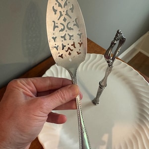 May include: A silver cake server with a decorative, openwork design is held above a white, tiered serving plate. The server has a long, ornate handle and a wide, slotted blade. The plate has a wavy, textured edge.