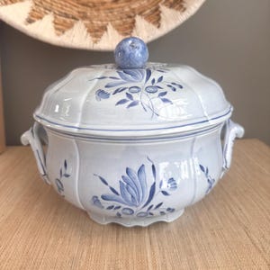 May include: A light blue ceramic tureen with a matching lid and two side handles. The piece features hand-painted dark blue floral motifs and foliage patterns. A round, blue-toned knob sits atop the lid, which has a scalloped edge design.