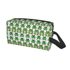 May include: Rectangular cosmetic bag with a repeating pattern of green cartoon frogs and small green hearts on a white background. The bag has a black zipper and a black handle.