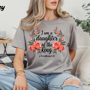 Puede incluir: Una camiseta gris con el texto "I am a daughter of the king" rodeado de un diseño floral con rosas rosas y hojas verdes. El texto "2 Corinthians 6:18" está debajo. La palabra "Grey" está en la esquina superior izquierda.