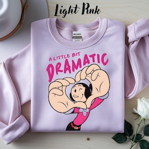 Puede incluir: Sudadera rosa claro con el texto "A Little Bit Dramatic" en rosa, con un personaje de dibujos animados formando un corazón con sus manos. La sudadera es de color pastel claro.