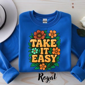 Puede incluir: Sudadera azul real con la frase "TAKE IT EASY" en letras naranjas y amarillas, rodeada de flores naranjas, verdes y amarillas. Un sombrero blanco y una rosa blanca en el fondo.