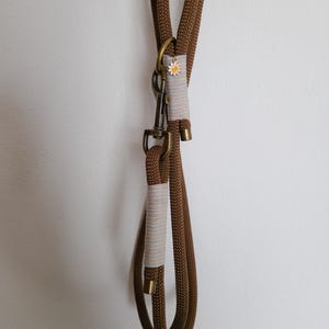 May include: A brown rope dog leash with a metal clasp and hardware. The leash has a decorative band with a small daisy accent. The leash is hanging against a white wall.