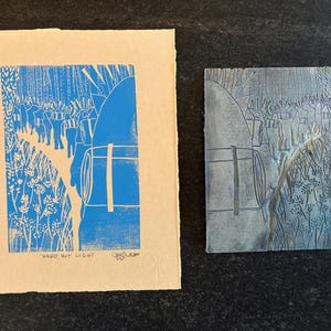 May include: A blue and white print of a landscape with figures and barrels, next to a gray relief print of the same scene. The print has the text "HARD, BUT LIGHT". The prints are on a beige background.