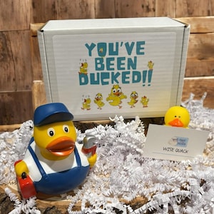 May include: A yellow rubber duck wearing a blue cap and overalls, holding a red tool box and a wrench. A white box with the text "YOU'VE BEEN DUCKED!!" and duck illustrations is behind the duck. A small card reads "WISE QUACK".