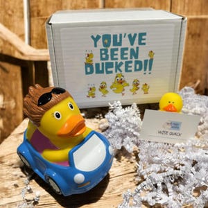 May include: A yellow rubber duck wearing sunglasses and a brown wig, driving a blue car. A small yellow duck sits on a white card that reads "Wise Quack". A white box with the text "YOU'VE BEEN DUCKED!!" is in the background.