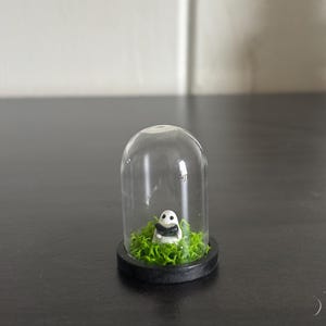 May include: A miniature panda figurine inside a clear glass dome. The panda is black and white, sitting on a bed of green moss. The dome has a black base and sits on a dark surface.