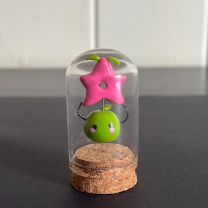 May include: A whimsical miniature sculpture inside a glass dome. The sculpture features a bright pink star above a green apple with rosy cheeks. The base is a cork, and the entire piece is set against a neutral background.