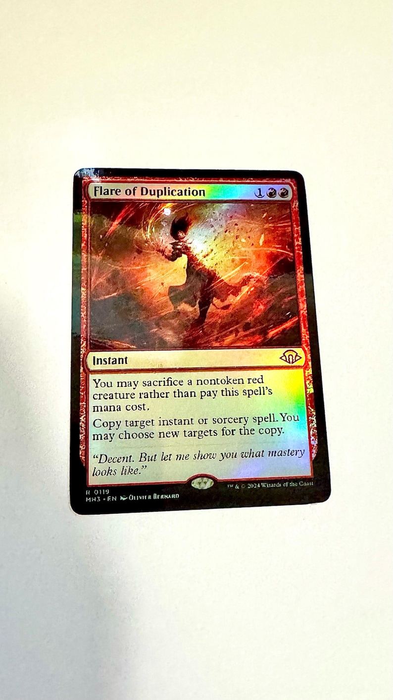 May include: A Magic: The Gathering card titled "Flare of Duplication." The card features a fiery, abstract illustration with red and orange hues. Text details the spell's effects, including copying instant or sorcery spells. The card is from the MH3 set.