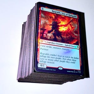 May include: A stack of Magic: The Gathering trading cards. The top card features the text "Rograkh, Son of Rohgahh" with a fiery background and a Kobold Warrior illustration. The card also includes the text "First strike, menace, trample Partner".