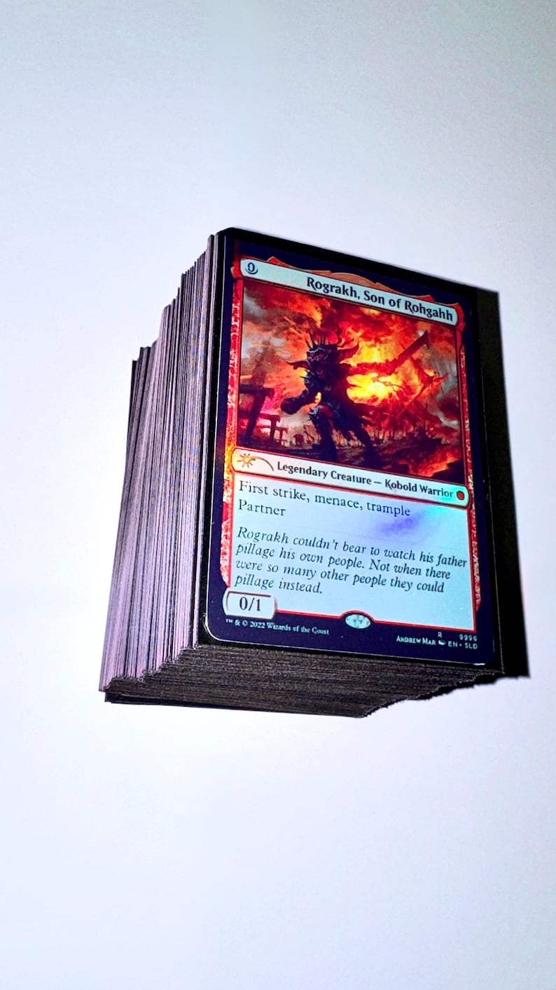 May include: A stack of Magic: The Gathering trading cards. The top card features the text "Rograkh, Son of Rohgahh" with a fiery background and a Kobold Warrior illustration. The card also includes the text "First strike, menace, trample Partner".