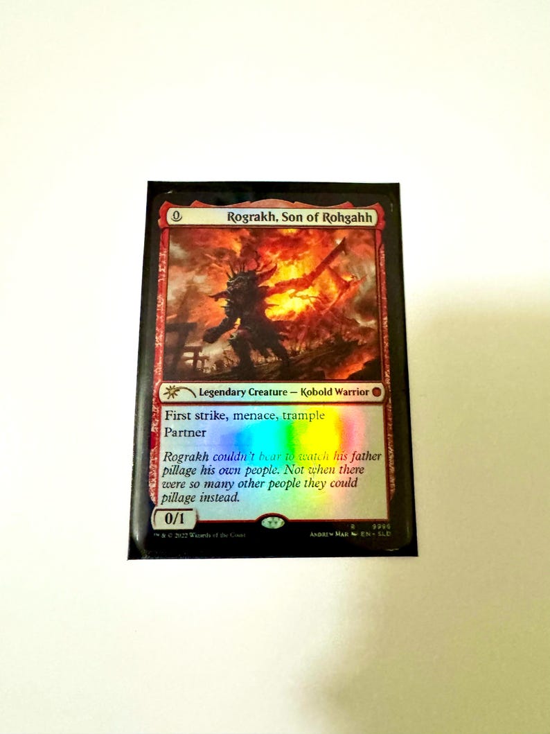 May include: A Magic: The Gathering card featuring Rograkh, Son of Rohgahh, a Kobold Warrior. The card has a fiery background with a silhouetted figure. Text includes "First strike, menace, trample, Partner" and flavor text. The card's power and toughness are 0/1.