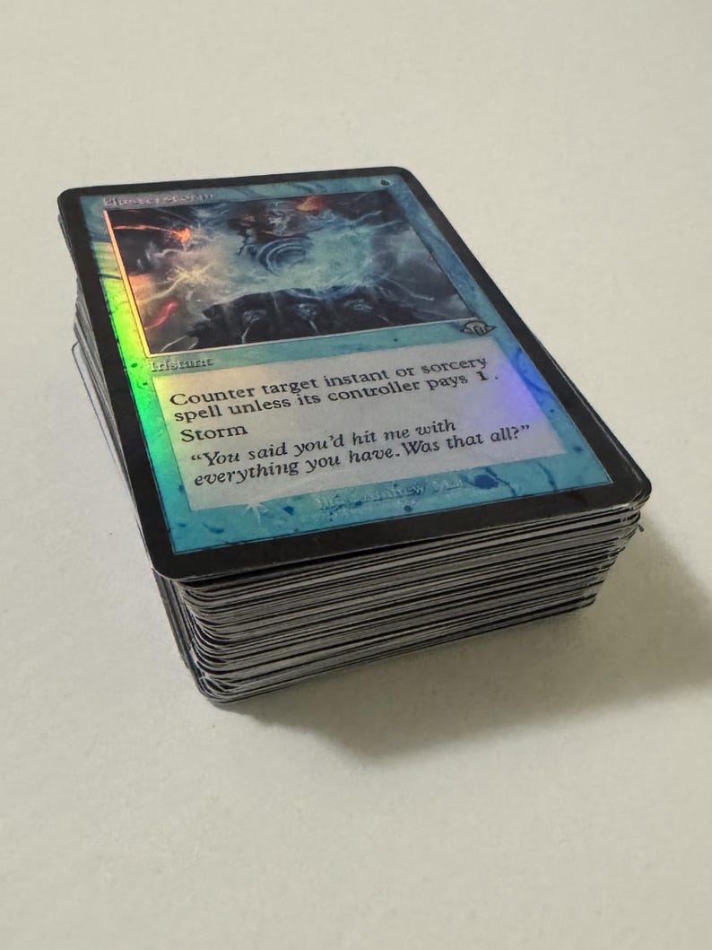 May include: A thick stack of trading cards featuring a holographic blue Magic: The Gathering card on top. The card is titled Flusterstorm and displays the text: Counter target instant or sorcery spell unless its controller pays 1. Storm.