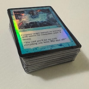 May include: A stack of Magic: The Gathering trading cards. The top card features a blue and holographic design with the text "Counter target instant or sorcery spell unless its controller pays 1. Storm". The card has a black border and a quote.