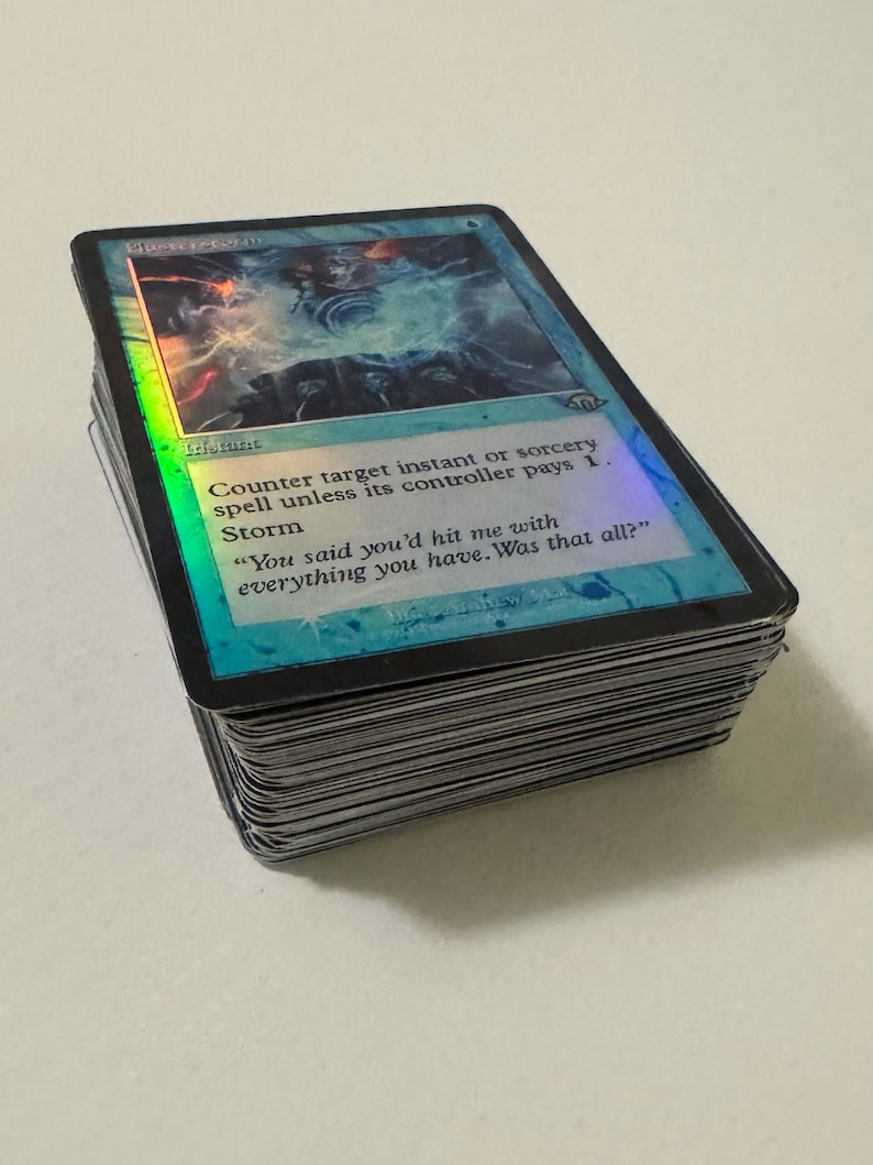 May include: A stack of Magic: The Gathering trading cards. The top card features a blue and holographic design with the text "Counter target instant or sorcery spell unless its controller pays 1. Storm". The card has a black border and a quote.