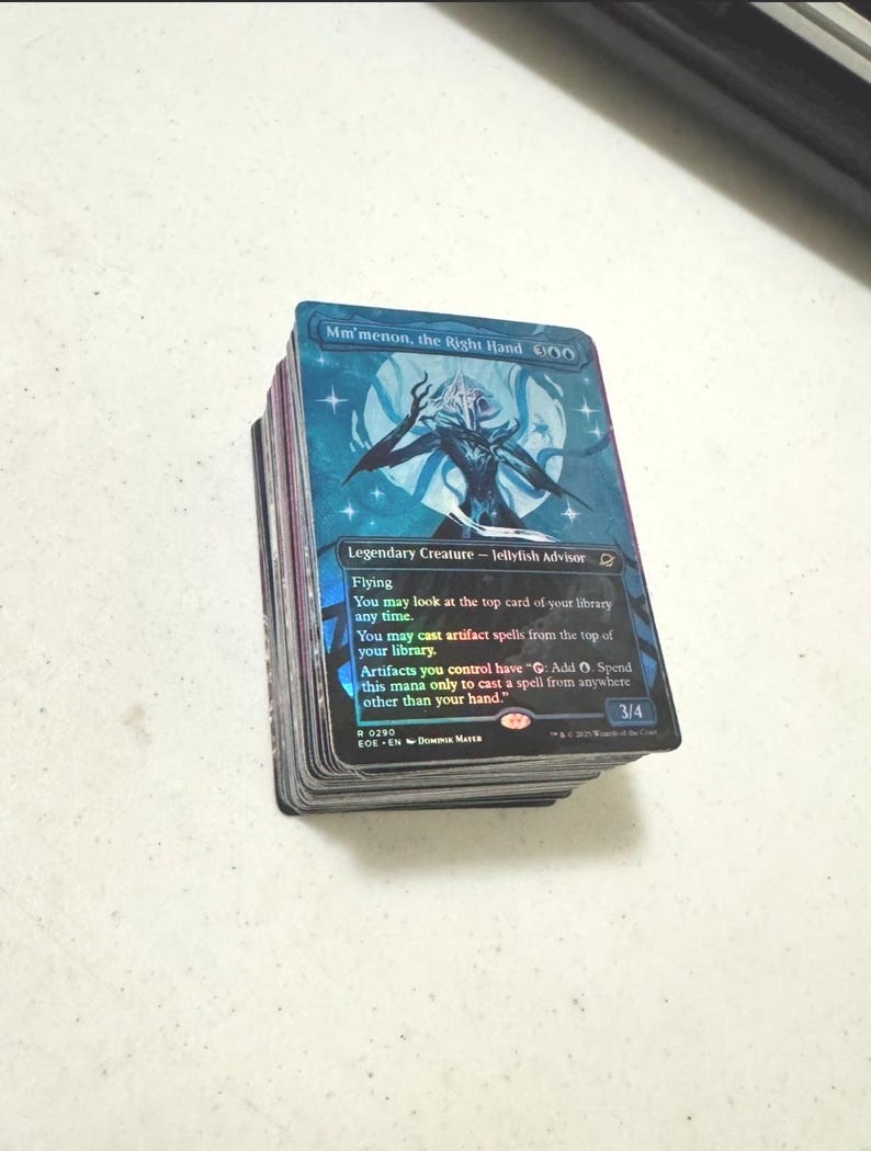 May include: A stack of trading cards featuring the holographic card Mm'menon, the Right Hand on top. The card displays a blue and purple illustration of a jellyfish-like creature, with text detailing its abilities as a Legendary Creature.
