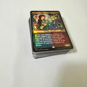 May include: A stack of Magic: The Gathering trading cards. The top card features an illustration of a woman in green and red, with the text "Jhoira, Ageless Innovator." The card has a holographic finish and includes game instructions.