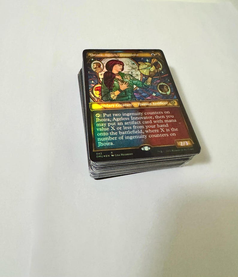 May include: A stack of Magic: The Gathering trading cards. The top card features an illustration of a woman in green and red, with the text "Jhoira, Ageless Innovator." The card has a holographic finish and includes game instructions.