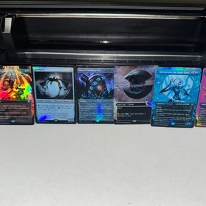 May include: A row of seven rectangular trading cards with holographic foil finishes, featuring various fantasy illustrations and printed text, arranged horizontally in front of a black electronic cutting machine on a plain white surface.