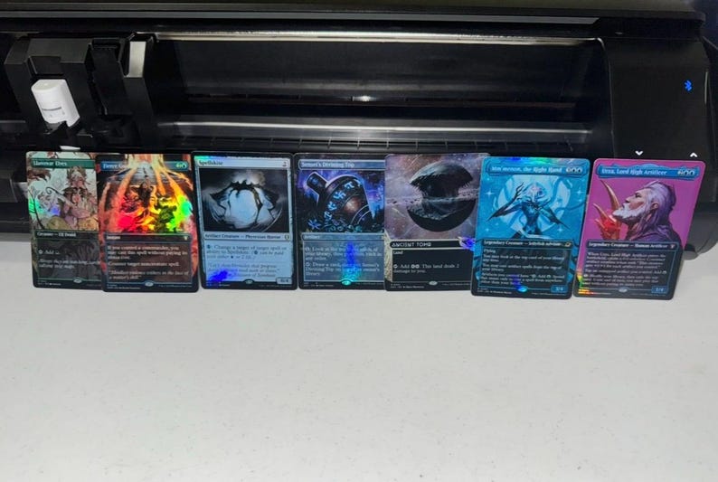 May include: A row of seven rectangular trading cards with holographic foil finishes, featuring various fantasy illustrations and printed text, arranged horizontally in front of a black electronic cutting machine on a plain white surface.