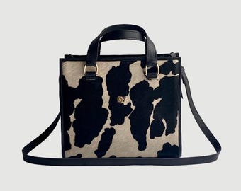 May include: A square handbag with a cowhide pattern in black and beige. The bag has black leather handles and a detachable shoulder strap. Gold-colored metal rings connect the handles to the bag.