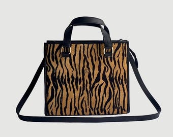 May include: A rectangular tote bag with a tiger stripe pattern in shades of brown and black. The bag has a black frame, handles, and a long, adjustable shoulder strap. Gold-colored hardware accents the handles.