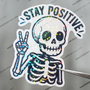 May include: A holographic sticker featuring a skeleton giving a peace sign. The sticker has the words "STAY POSITIVE" in a curved font above the skull. The design has a glittery, iridescent effect, with a silver and black color scheme.