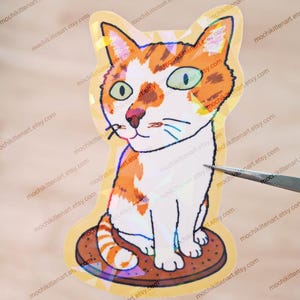 Op de afbeelding: Een gestanste sticker met een illustratie van een wit-oranje cyperse kat die op een bruin rond koekje zit. De kat heeft grote groene ogen en een roze neus. De sticker heeft een holografische afwerking die licht reflecteert in patronen.