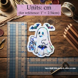 May include: A holographic sticker featuring a white ghost holding a lantern, with two small bats above and one bat below. The sticker is positioned next to a clear plastic ruler showing a height of approximately 5 cm and a width of 5 cm.