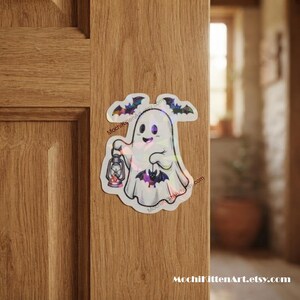 May include: A holographic sticker featuring a white ghost holding a lantern and a small bat, with two flying bats above. The text MochiKittenArt.etsy.com is printed in small black letters along the side of the ghost figure.