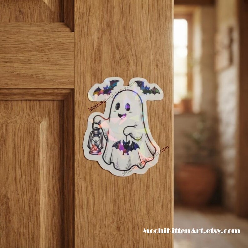 May include: A holographic sticker featuring a white ghost holding a lantern and a small bat, with two flying bats above. The text MochiKittenArt.etsy.com is printed in small black letters along the side of the ghost figure.