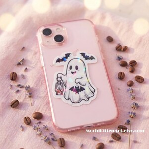 May include: A light pink smartphone case featuring a holographic sticker of a white ghost holding a lantern and a small bat, with additional bats flying above. The sticker has a glossy finish that reflects light in various colors.