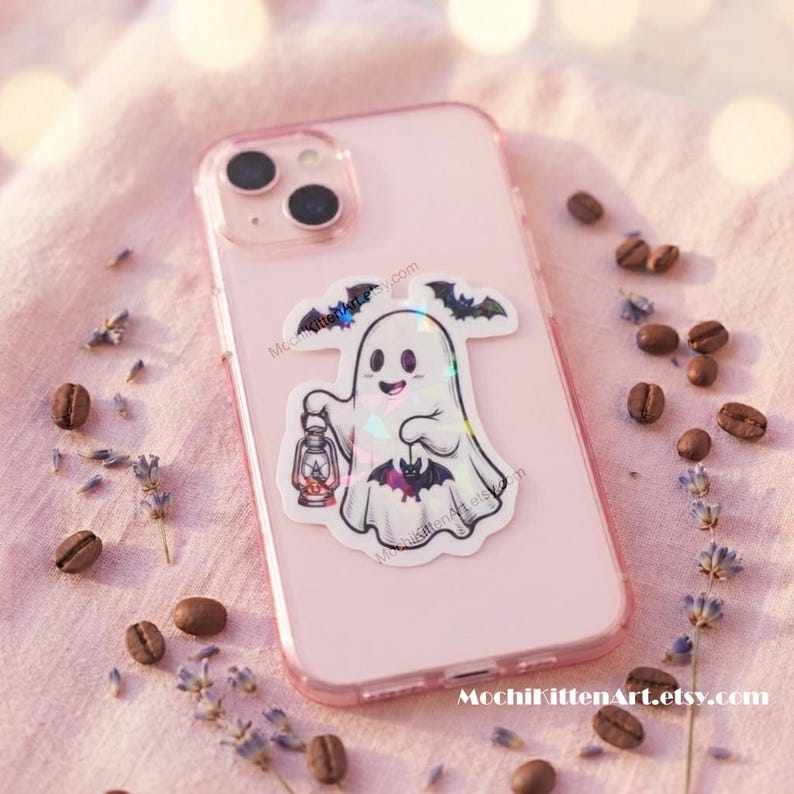 May include: A light pink smartphone case featuring a holographic sticker of a white ghost holding a lantern and a small bat, with additional bats flying above. The sticker has a glossy finish that reflects light in various colors.