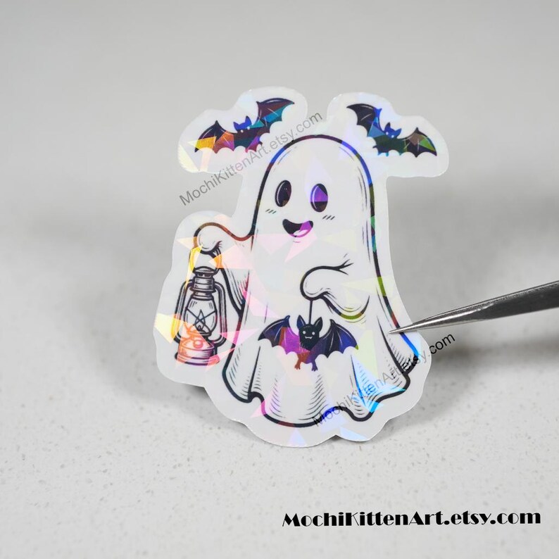 May include: A holographic sticker featuring a white ghost with a smiling expression, holding a lantern in one hand and a small purple bat in the other. Two additional bats are positioned above the ghost. The text MochiKittenArt.etsy.com is visible.