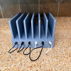 May include: A light blue multi-device charging station featuring six vertical slots for organizing electronics. The base includes six integrated ports with four black charging cables and two white charging cables plugged into the front panel.